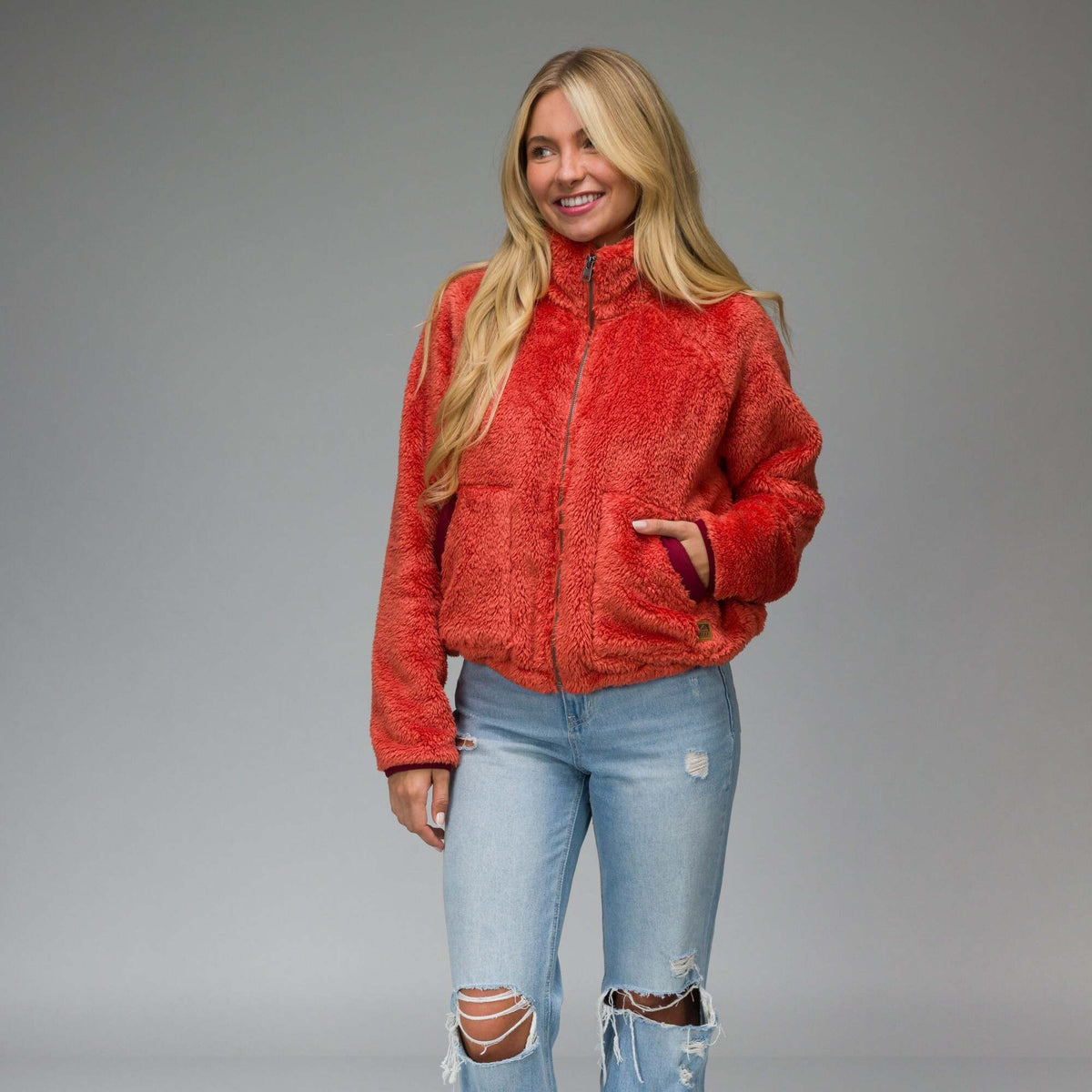 Woman wearing a bright chili red Pilot Sherpa Jacket facing front with hands in pockets, showing its cozy textured fabric.