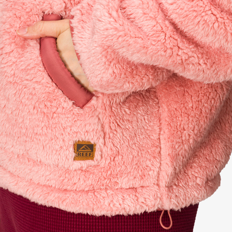 Close-up of a woman’s hand in the front pocket of a coral pink Pilot Sherpa Jacket showing soft, cozy texture and REEF logo.