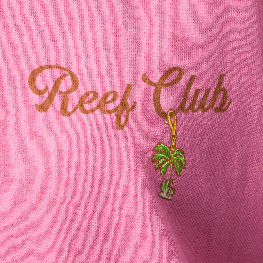 Close-up of pink Haus Boyfriend Crew fabric showing "Reef Club" text and embroidered palm tree charm, surf-inspired comfort.
