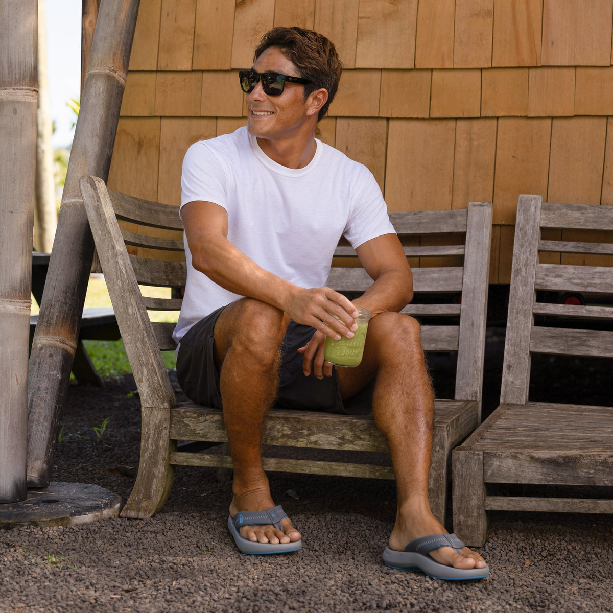 Man wearing Cruiser sandals facing forward, seated on a wooden bench outdoors, holding a green drink.