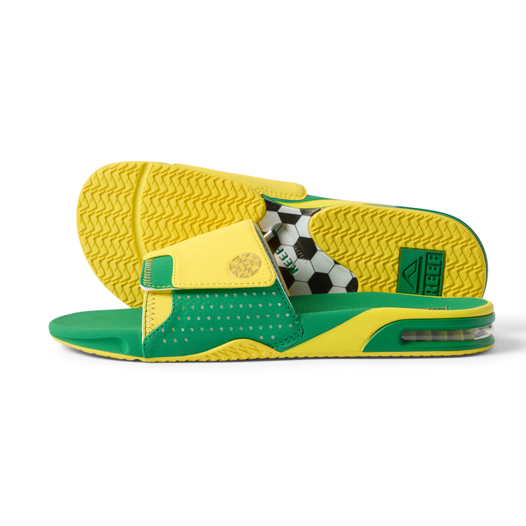 Pair of Fanning Slide sandals facing right and sole up, men's green and yellow water-friendly slides with cushioned EVA footbed and soccer ball graphic underfoot.