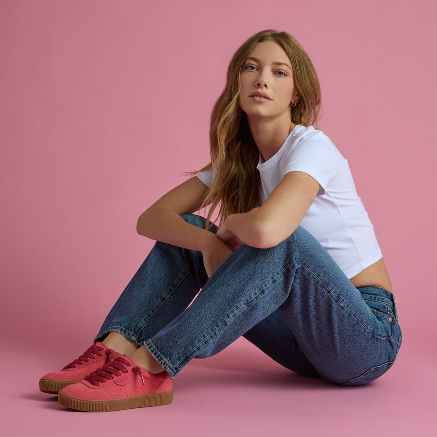 Woman seated against pink backdrop wearing white top, jeans, and red Lay Day Seas shoes facing left, highlighting comfort and style.