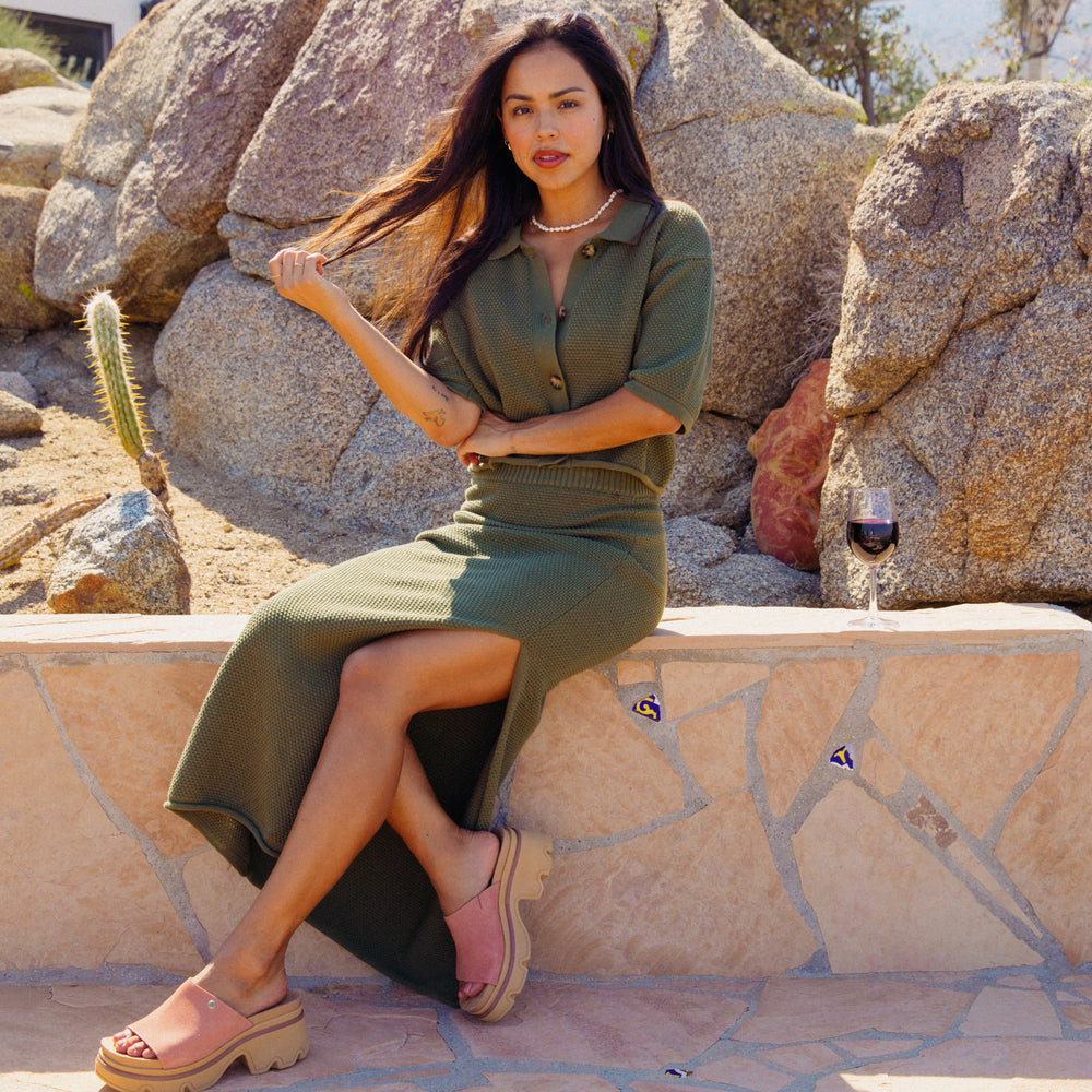 Woman wearing olive Up North Sweater Top seated outdoors showing front and side view with pink comfort sandals facing right