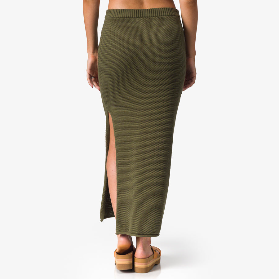 Back view of woman wearing olive Up North Sweater Skirt with side slit, paired with surf-inspired sandals facing forward.
