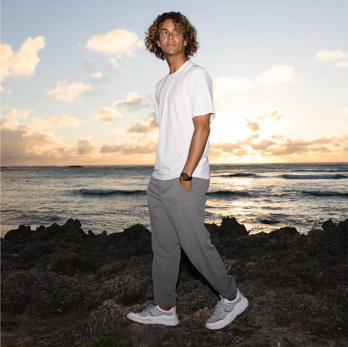 Man wearing Shorebreak TRV shoes facing left in grey walking pants, standing on rocky beach at sunset with ocean background.