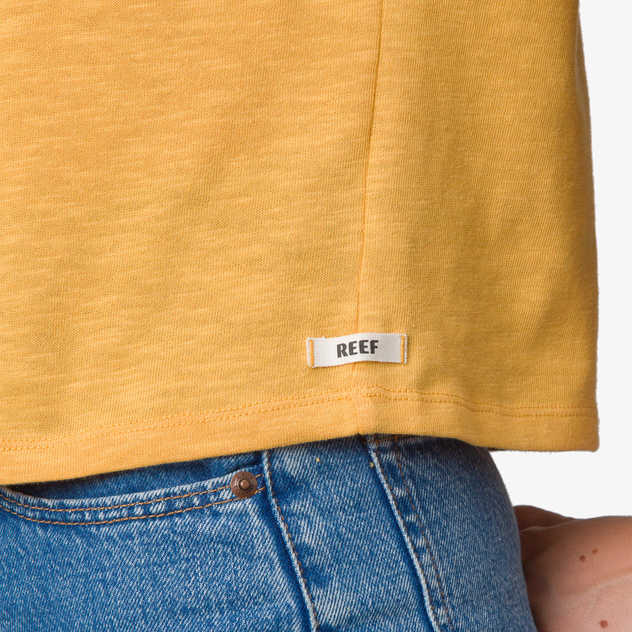 Close-up of the bottom hem of the Ringer Tee in honey gold showing soft fabric and REEF label above blue denim jeans.