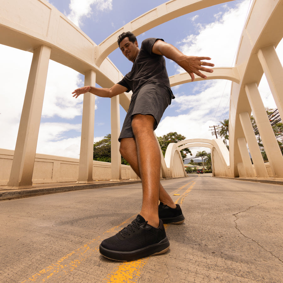 Man wearing Shoreline Del Mar black sneakers facing forward on bridge, showcasing durable, comfortable beach-inspired footwear.