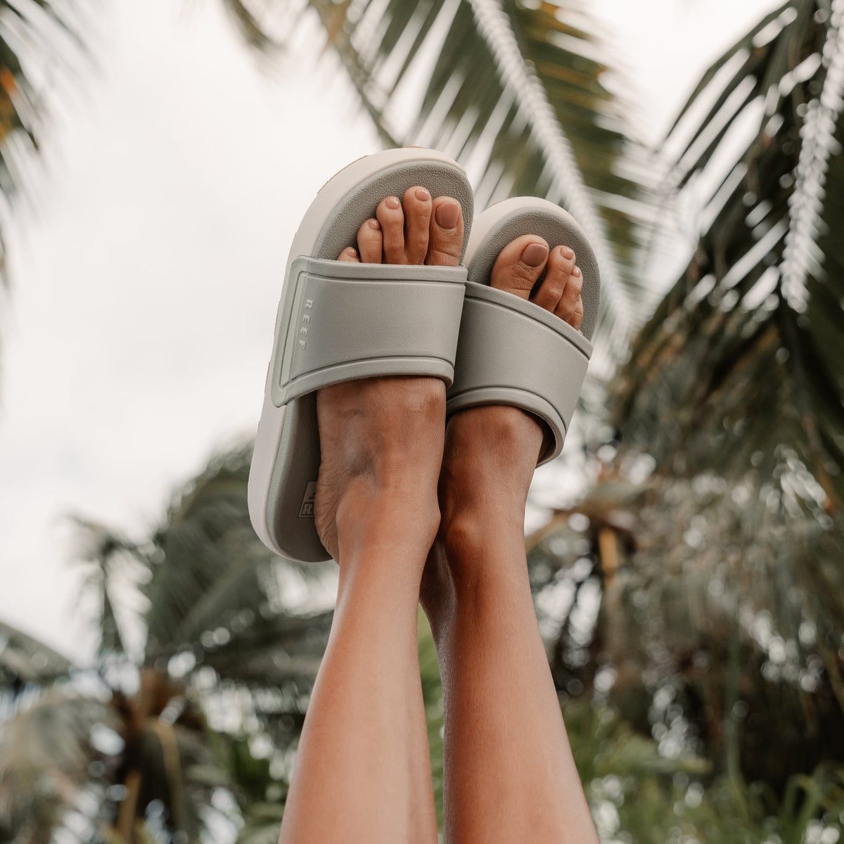 Sydnee Cutback Low - Sage - on a pair of feet up in the air with palm trees in the background