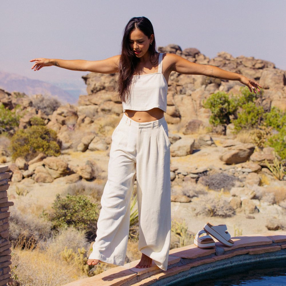 Woman wearing Tori Linen Tank in almond milk, standing barefoot near pool with arms outstretched, desert background