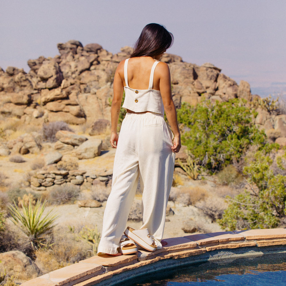 Woman wearing Tori Linen Tank in almond milk facing away, standing by pool with desert rocks in background