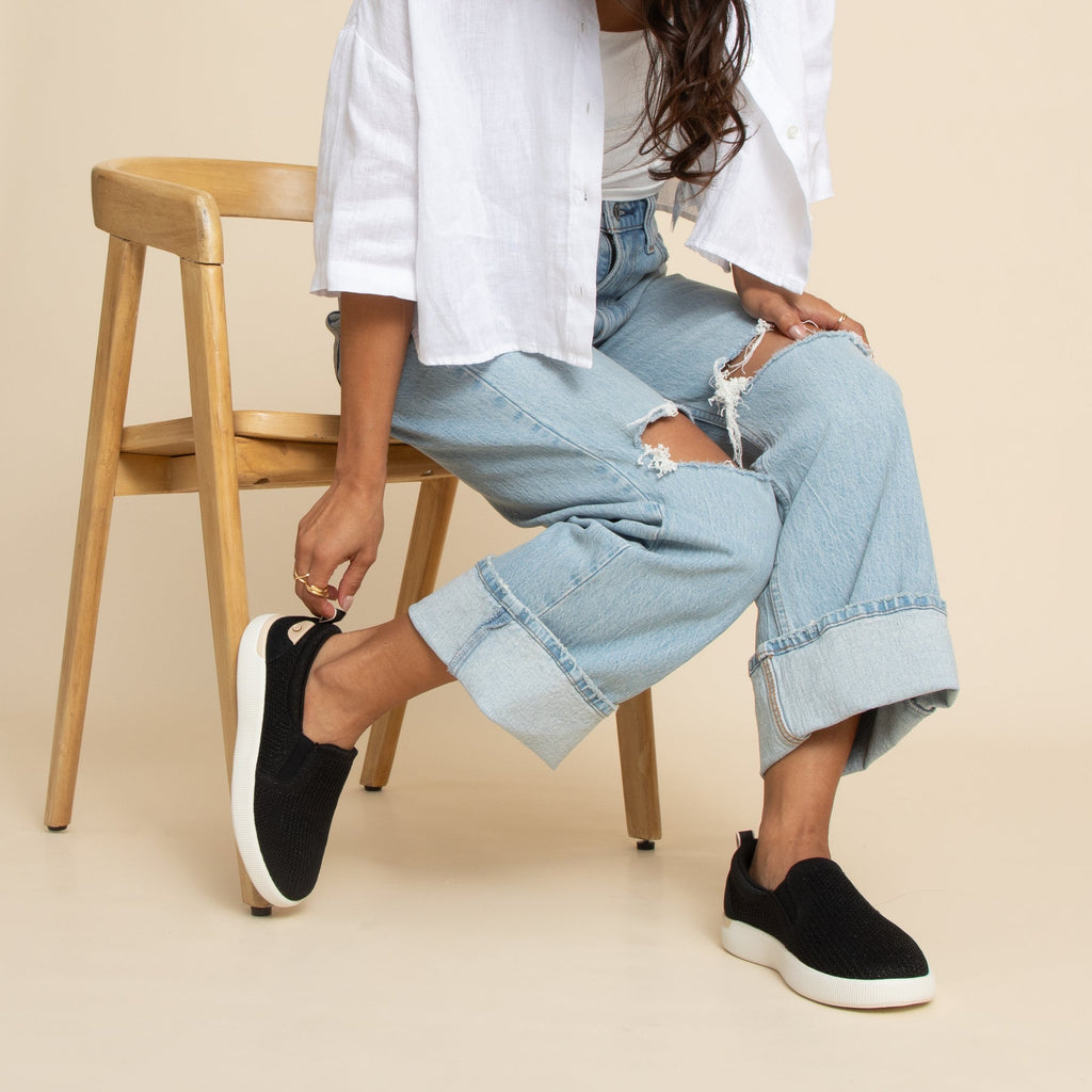 Neptune Elyse black slip-on shoes facing right with soft cushioning and anatomical arch support on model sitting on wooden chair